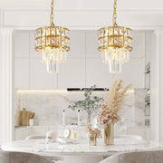 A luxurious kitchen features two elegant Gio Crystal Kitchen Pendant Lights by Morsale, gracefully hanging over a round marble table. The table is adorned with decorative vases, a clock, and candlesticks. In the background, modern style shines through with white cabinets and a marble backsplash.