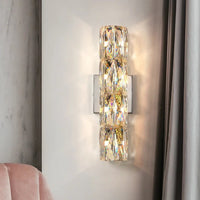 The Morsale Bacci Crystal Wall Sconce, featuring multiple faceted panels, emits warm light on a white wall beside a gray curtain, with part of a light pink chair in the corner.