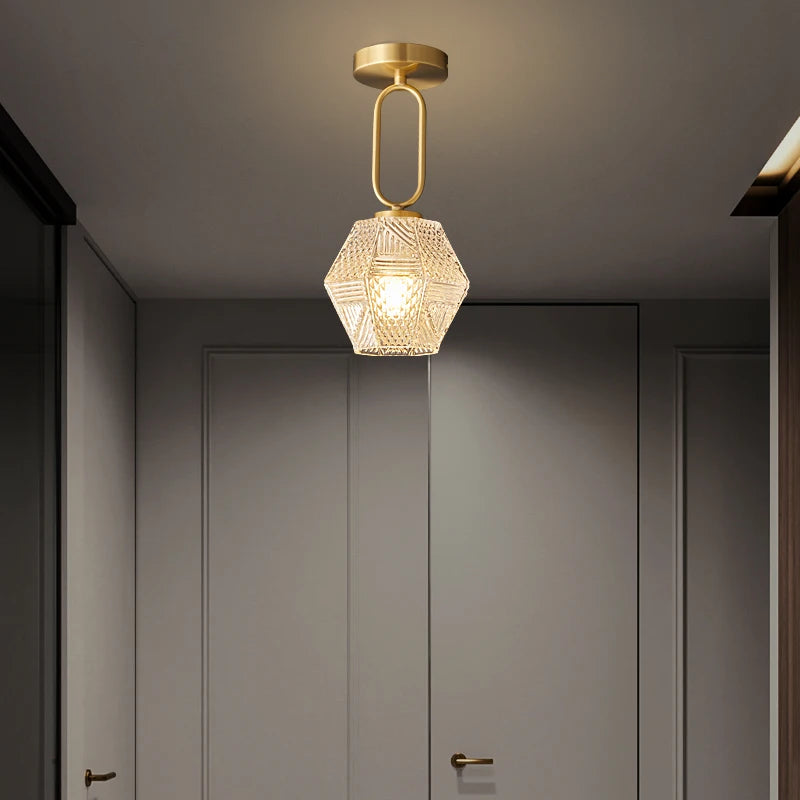 A modern hallway with a closed door showcases the Palermo Ceiling Light by Morsale, featuring LED bulbs. The pendant has a hexagonal, textured glass shade and a gold-colored fixture, casting a warm glow and enhancing the modern style of the space.