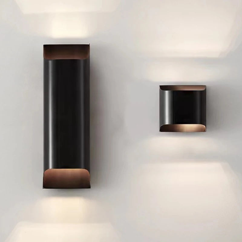 The Rame Copper Sconce by Morsale features minimalist LED wall lamps in a modern design. The left lamp is an elongated rectangle with soft illumination, while the right is a smaller square. Both lamps have a sleek black finish, emitting warm light from the top and bottom, and are subtly enhanced by brass details for added elegance.