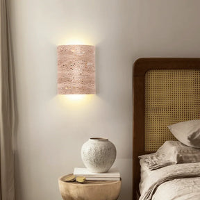 A Natural Travertine Art Deco Wall Lamp from Morsale.com, crafted from beige, porous stone and shaped into a cylinder, is mounted on a white wall. The light radiates softly from both the top and bottom, generating an ambient glow around the fixture that accentuates its ART DECO design.