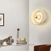 A modern interior space with a minimalistic design. The Menards Modern Marble Wall Lamp by Morsale, featuring LED bulbs, emits a circular, glowing light on the wall. Below, a table holds an abstract sculpture and a plate with cheese. A framed artwork is partially visible on the wall to the left.
