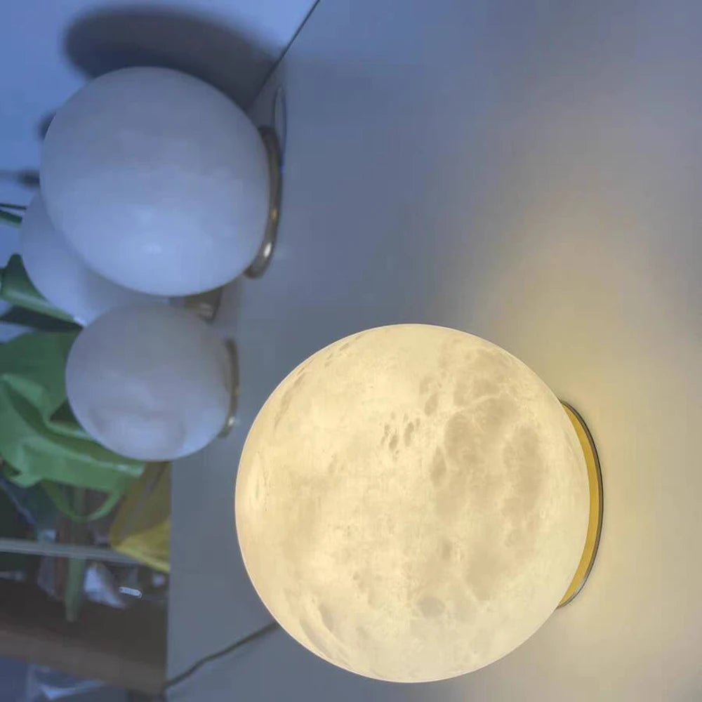 The Moonshade Marble Ball Light Fixture by Morsale emits a soft glow on a reflective surface. In the background, marble lamps add modern style lighting, while two round white objects enhance the composition's depth and serenity.