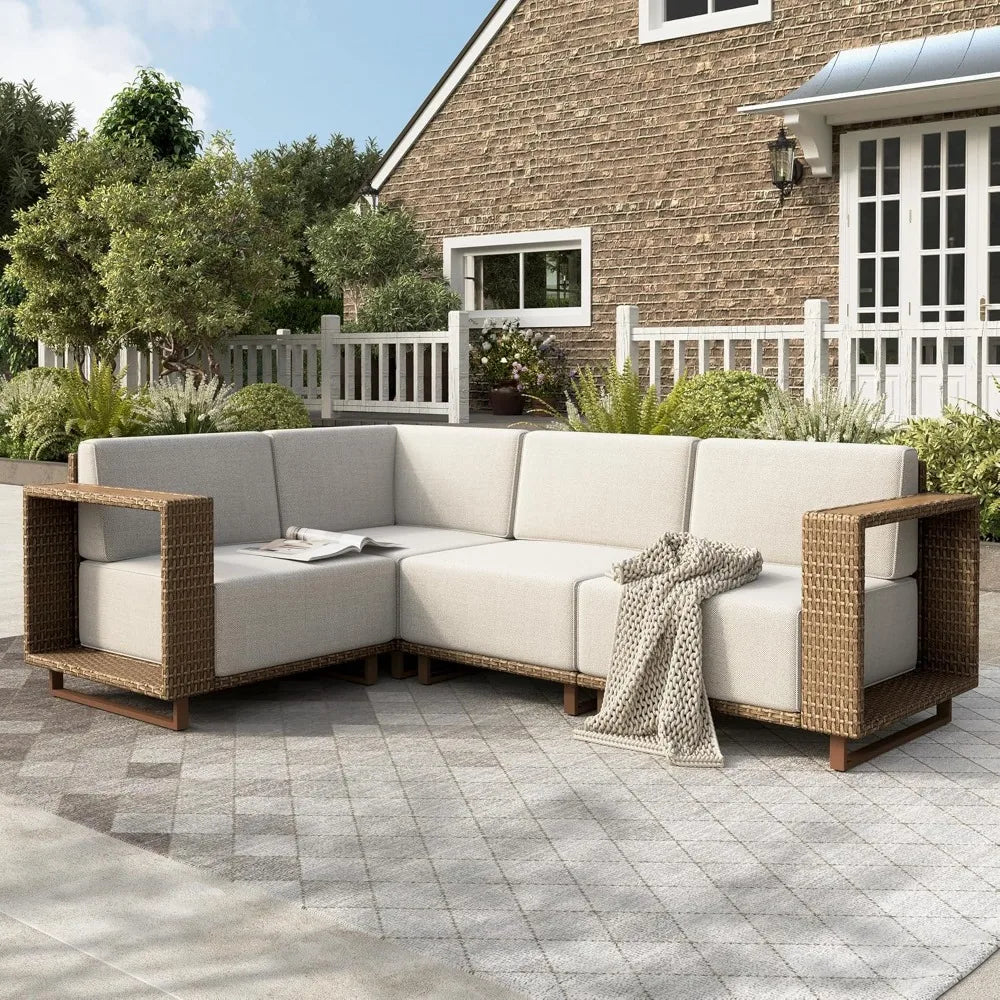 An outdoor patio features the modern Morsale Burton Outdoor Rattan Wicker Sofa Sectional with beige cushions and a knitted throw. It sits on a patterned tile patio surrounded by greenery, in front of a brick house with white trim and a picket fence.