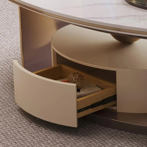 The Morsale 55" Modern Cocktail Table is round and beige, featuring a high-temperature resistant marble top and a partially opened wooden drawer containing a small cube puzzle and a closed book. It sits elegantly on a gray textured carpet.