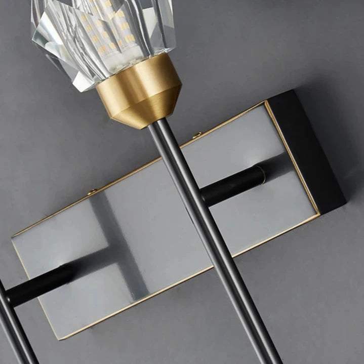 A close-up of a modern wall-mounted Morsale Diamante Crystal Wall Sconce highlights its geometric design, LED lighting, and a metallic base with black and gold accents. The faceted clear glass bulb cover stands out beautifully against the gray background, making it an elegant addition to home decor.