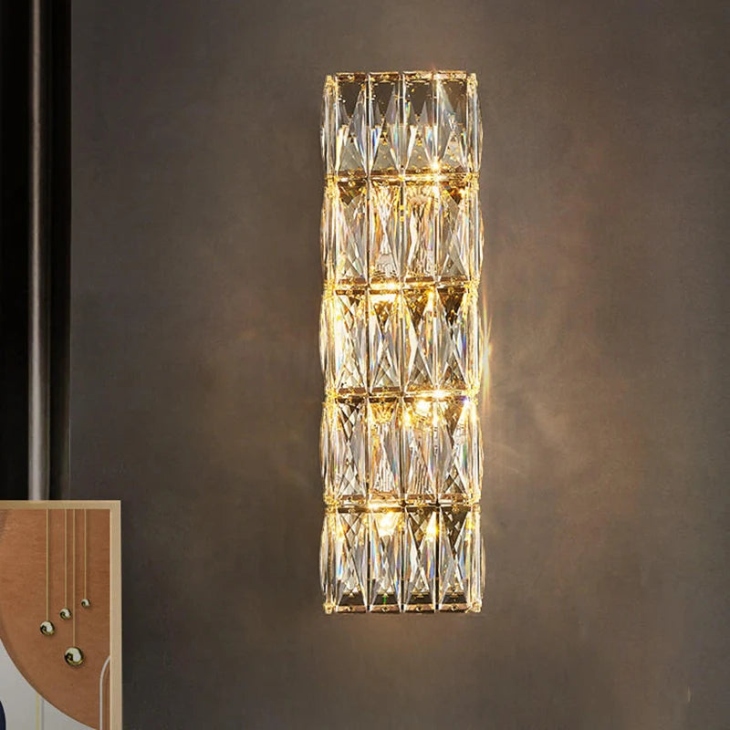 A Bacci Crystal Wall Sconce by Morsale, showcasing a vertical rectangular design in a modern style, casts a warm, golden light against the dark gray wall. Its geometric design with faceted surfaces reflects the glow of the LED bulb, creating a soft ambiance. A framed artwork is partially visible nearby.