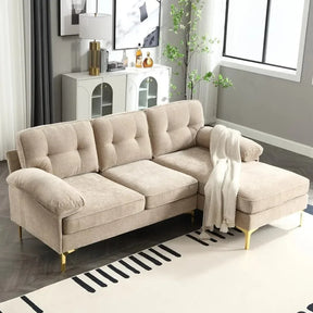The bright living room is anchored by the Morsale 83" Sectional Sofa with Chaise, featuring beige tufted cushions and gold legs. A white throw blanket adds coziness, complemented by a decorative plant, side cabinet, and a large window with gray curtains.