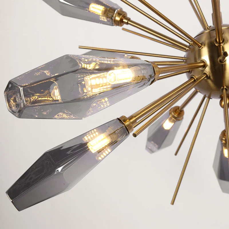 A close-up of the Morelli Sputnik Chandelier by Morsale.com featuring a gold, spiked central hub from which elongated, geometric glass shades extend. Each shade has a visible LED bulb inside, emitting warm light. The design is sleek and contemporary with a touch of elegance and is fully dimmable for any mood.