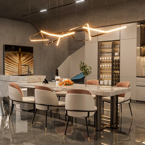 A modern kitchen features a large marble island surrounded by three sets of Vegan Leather Dining Chairs by Morsale. A contemporary abstract light fixture hangs overhead, with sleek cabinets, a display shelf with jars, and minimalist wall art in the background.
