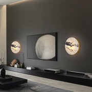 A modern living room with a dark wall showcases a large circular artwork, flanked by two Morsale Moonshade Natural Marble Sconce Lights. The Art Deco-inspired space features a low black entertainment unit and minimalist decor.