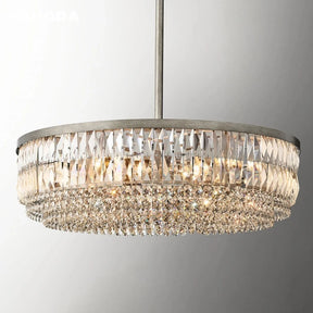 A close-up view of the Bacci Crystal Chandelier by Morsale.com reveals multiple tiers of K9 crystals. The chandelier features a circular, luxurious gold finish frame and emits a warm, glittering light that casts beautiful reflections from the intricate crystal detailing against a plain, light grey wall.