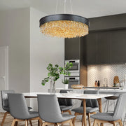 A modern dining room features a wooden table surrounded by gray chairs, illuminated by the Alleri Modern Chandelier from Morsale, designed in black and gold. A potted plant sits at the center of the table, with a kitchen in the background showcasing dark cabinets and a herringbone white backsplash that epitomizes modern style.