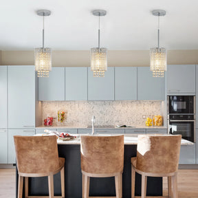 A modern kitchen features Morsale's Alleri Crystal Pendant Lights, Set of 3, above a marble island with four beige bar stools. Light gray cabinets and a marbled backsplash enrich the sleek design. Colorful jars and a built-in oven add vibrant touches to the space.