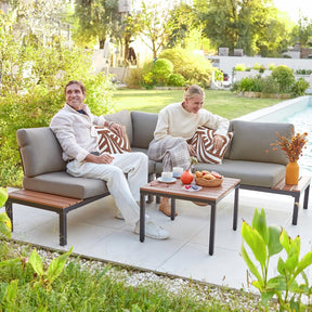 A man and woman sit on a Morsale Warton Patio Set, smiling and relaxed. The setting includes a table with drinks, pastries, and flowers in a sunny garden by a pool, surrounded by greenery and decorative pillows.