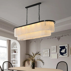 A modern dining room features the Moonshade Marble Chandelier by Morsale with LED bulbs, its cascading fabric layers lighting an oval table for four. Decorative molding, framed abstract art, and natural light through a tall window enhance the space's modern style.