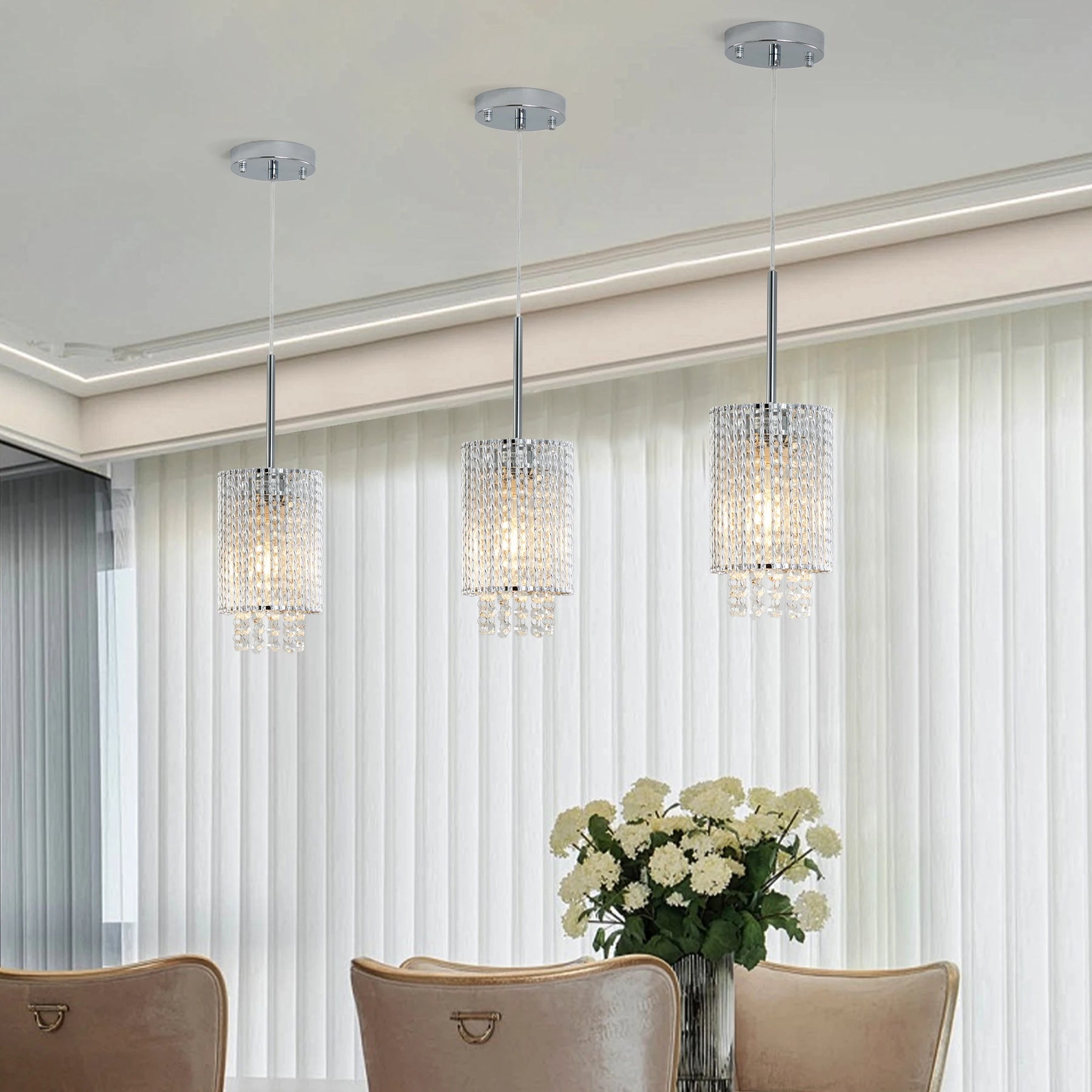 A modern dining room showcases premium materials with Morsale's Alleri Crystal Pendant Lights, Set Of 3 hanging elegantly from the ceiling. Beige chairs encircle a round table with a vase of white flowers, while vertical blinds drape the tall windows.