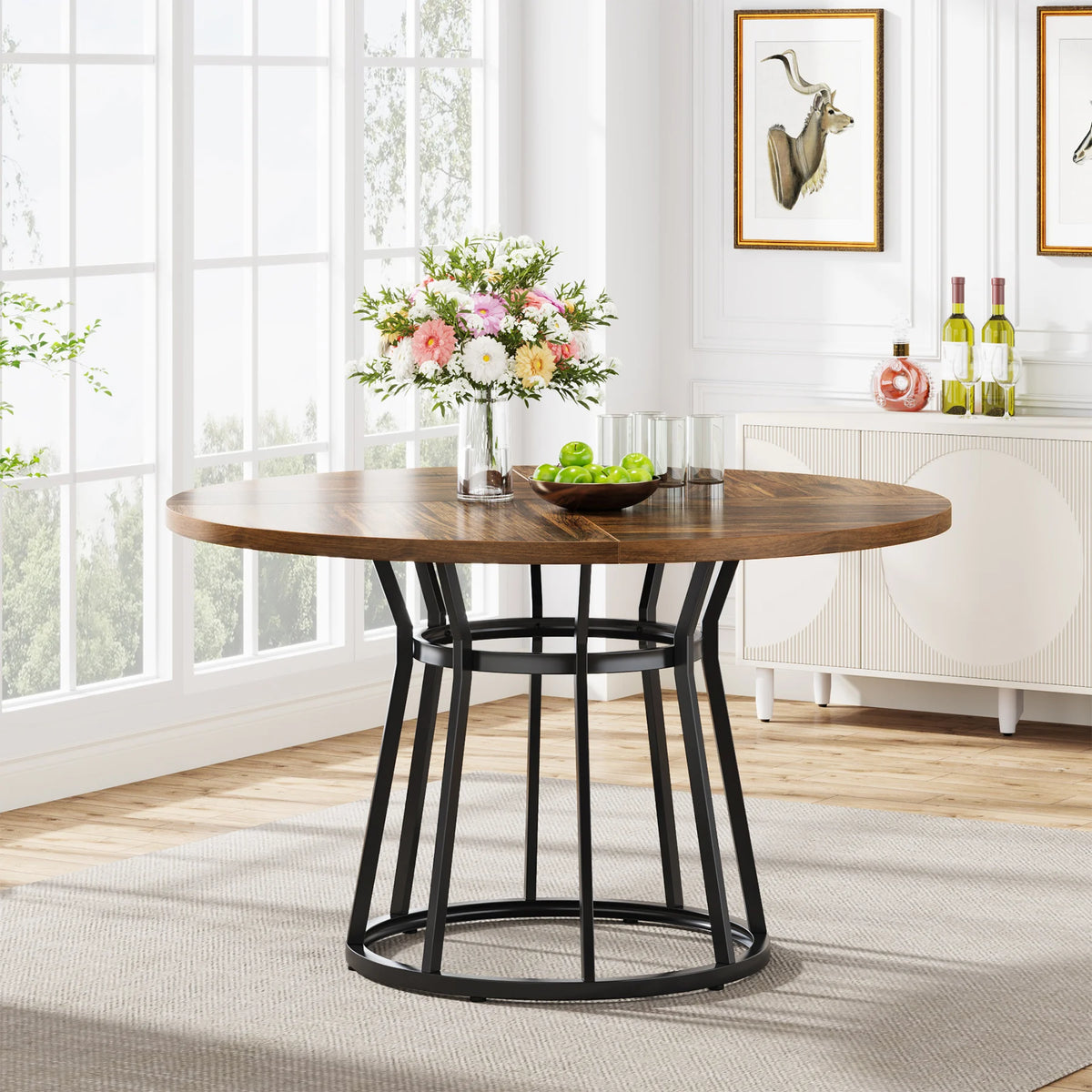 The Morsale Bradford 47-Inch Round Kitchen Dining Table, featuring a black metal frame and wooden top, exudes modern style on a gray rug. A colorful vase and green apples decorate it as natural light highlights art and wine bottles on the sideboard against large windows.
