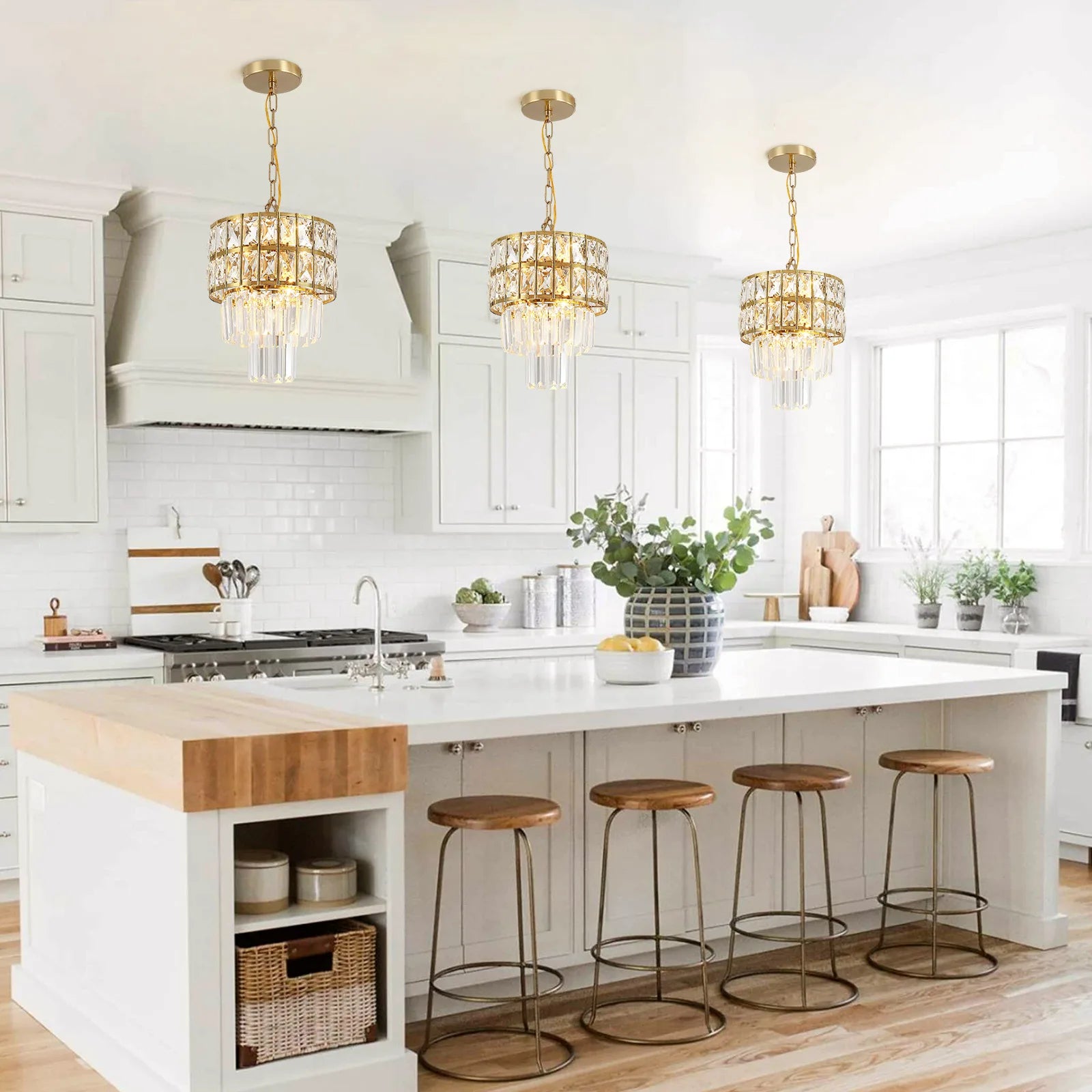 A bright, modern kitchen with white cabinets showcases a large island accented with wood and four wooden stools. The contemporary style is elegantly enhanced by three Morsale Gio Crystal Kitchen Pendant Lights hanging above. Natural light streams in through large windows, illuminating the counter where a vase with green foliage is displayed.