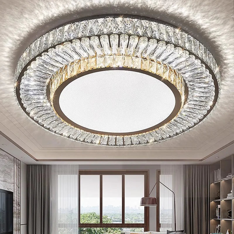 The Morsale Gio Crystal Ceiling Chandelier, adorned with crystal design elements and LED bulbs, is elegantly mounted on the ceiling. Below, a modern living room showcases a large window with curtains, a floor lamp, and shelves. Natural light enhances the gentle ambiance of the space.