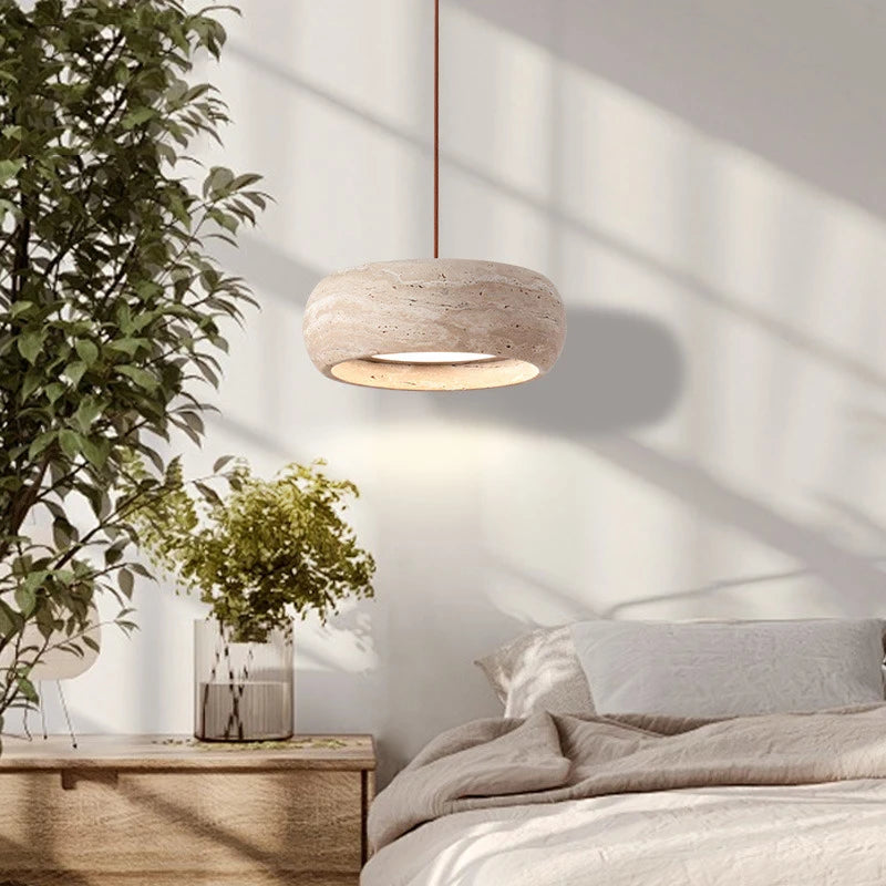 A cozy bedroom features a soft beige bed and Morsale's Modern Travertine Pendant Lights For Kitchen above. Sunlight streams through a window, casting shadows on the wall, while a bedside table with greenery in a vase adds a natural touch.