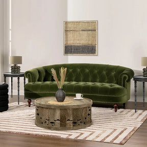 A living room includes a Chesterfield Velvet Tufted Sofa in Olive Green by Morsale, a round wooden coffee table, two side tables with matching lamps, and is adorned with a neutral striped rug. A textured abstract art piece enhances the elegance of the light-colored walls.