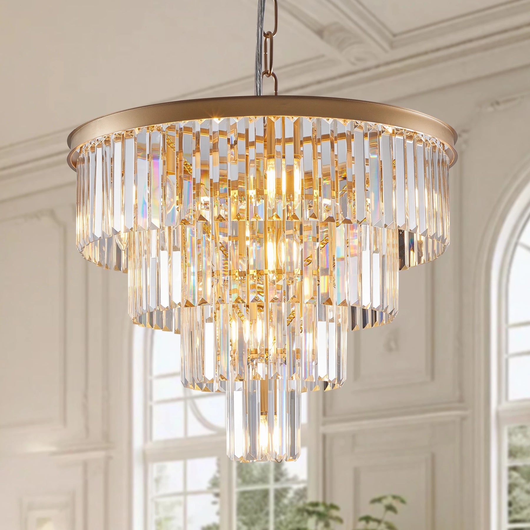 The 23.6" Gio Crystal Chandelier by Morsale, with its luxurious gold frame and cascading crystal prisms, hangs from the ceiling in a bright contemporary interior, complementing tall arched windows in the background.