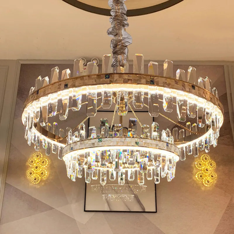 The Calvi Crystal Chandelier by Morsale is a luxurious LED lighting fixture featuring two circular tiers and adorned with hanging crystal prisms, casting a warm and elegant glow. It complements a setting with soft beige walls and subtle decorative patterns.