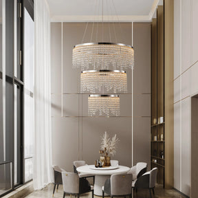 A modern dining room with two Morsale Alleri 3-Tier Crystal Chandeliers and sleek pendant lights includes a round table surrounded by gray chairs. Tall windows bring in natural light, reflecting off decorative vases on the table illuminated by energy-efficient LED bulbs.