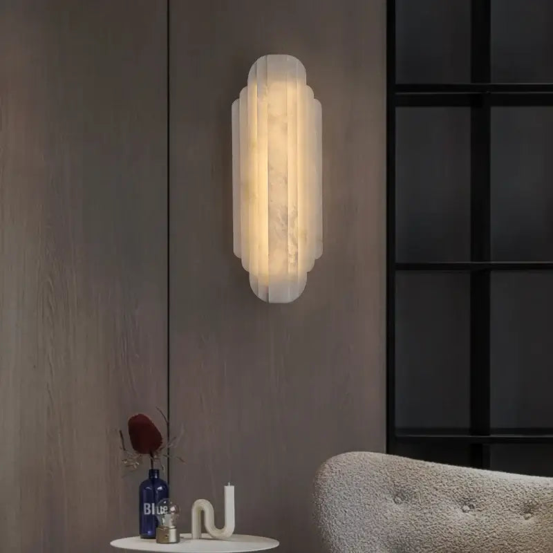 
                  
                    The Morsale Arina Marble Sconce features a frosted glass design and emits warm light on a wooden panel wall. Beneath it, a small table displays a blue bottle, décor piece, and part of a gray upholstered chair is visible.
                  
                
