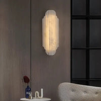 The Morsale Arina Marble Sconce features a frosted glass design and emits warm light on a wooden panel wall. Beneath it, a small table displays a blue bottle, décor piece, and part of a gray upholstered chair is visible.