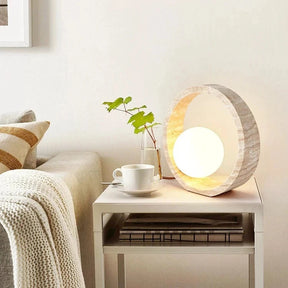 The Modern Travertine LED Table Lamp by Morsale.com features a round Natural Yellow Cave Stone frame with a textured, natural finish and a glowing spherical light bulb in the center. The lamp emits a warm, ambient light, creating a cozy atmosphere, and has a cord extending from the back.