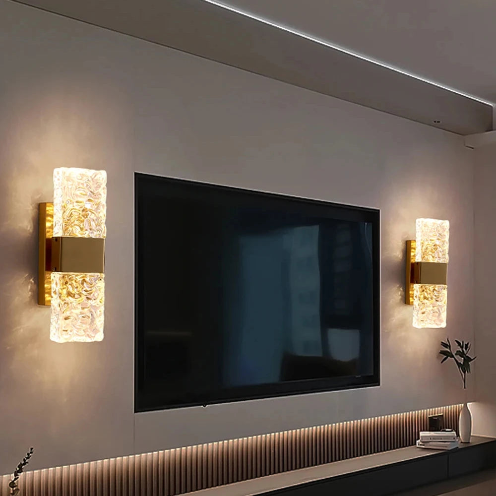 A modern living room showcases a large flat-screen TV mounted on the wall, accompanied by two Morsale Water Ripple Crystal Wall Sconces with textured glass and touch switches that emit a warm glow. Beneath the TV is a cabinet featuring a subtle wood pattern and stainless steel accents, enhanced by the presence of a small plant.