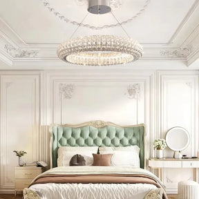 An elegant bedroom featuring a plush, tufted mint-green headboard and ornate white walls is illuminated by the Bacci Crystal Modern Chandelier from Morsale. A white and beige bedspread complements the decor, while a vanity with a round mirror and small plants adds to the contemporary style.