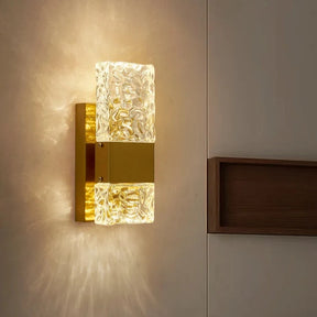 The Morsale Water Ripple Crystal Wall Sconce, featuring a golden base and textured glass cover, radiates warm, ambient light. It is installed on a neutral-colored wall beside a wooden shelf. The touch switch allows for effortless control of the soft glow and intricate patterns it projects onto the surrounding surfaces.