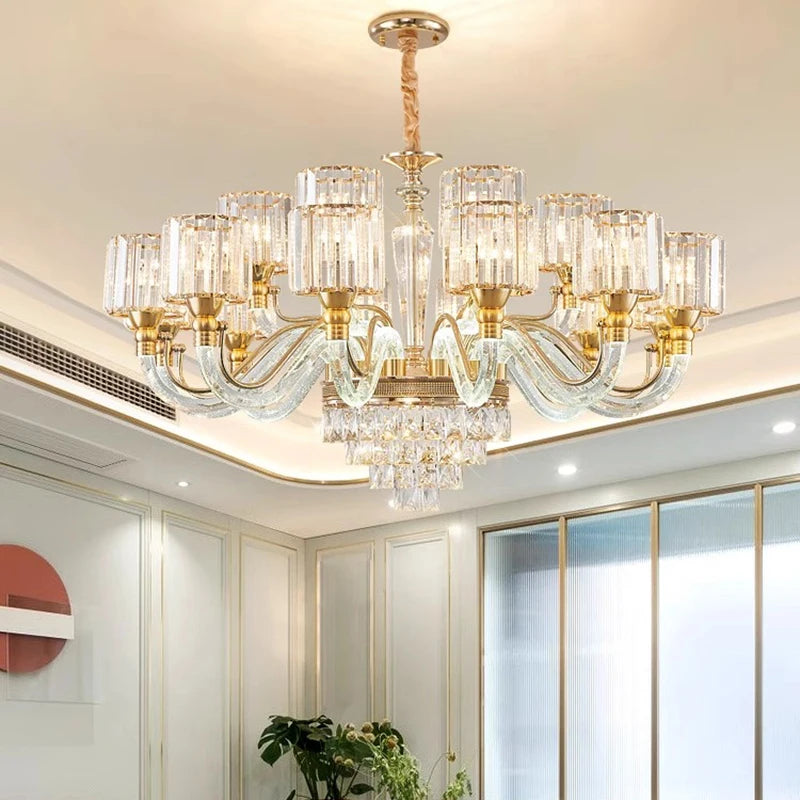 A Gio Crystal Contemporary Chandelier by Morsale, featuring gold accents, crystal details, and multiple glass light fixtures with LED bulbs, elegantly hangs from the ceiling in a modern room with light-colored walls. In the background, a round decorative wall piece can be seen.