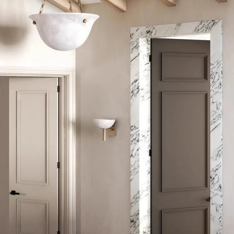 A modern interior showcases a marble-framed doorway with a partially open beige door, adjacent to another beige door on the wall. The space is illuminated by contemporary lighting, including a Morsale Spanish Marble & Copper Antique Sconce and a white bowl-shaped ceiling light.