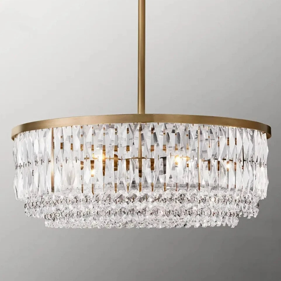 The Bacci Crystal Chandelier by Morsale.com boasts a luxurious gold finish and intricate, cascading K9 crystals that sparkle. The cylindrical chandelier features multiple layers of crystal drops gracefully hanging from a central rod, set against a light gray background.