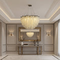 A modern bedroom exudes elegance with the Alleri Crystal Pendant Chandelier by Morsale.com hanging above a neatly made bed, featuring green and neutral bedding, a dark green accent wall, and sheer white curtains letting in natural light.