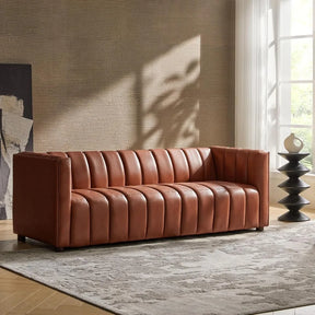 The Biagio 83" Genuine Leather Sofa by Morsale with a ribbed design is placed on a textured gray rug in the modern living room. Sunlight filters through sheer curtains into a large window, casting a warm glow. A decorative vase adorns a stacked black side table, enhancing the mid-century vibe.