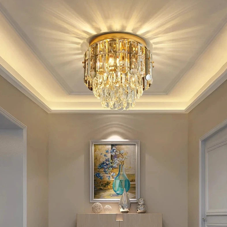 A gold Giano Ceiling Light Fixture from Morsale.com with handmade crystals hangs from the ceiling in an elegantly decorated room. The fixture's light creates a radiant pattern on the ceiling. Below the chandelier is a console table with decorative items, and a painting of a vase with flowers is displayed on the wall.