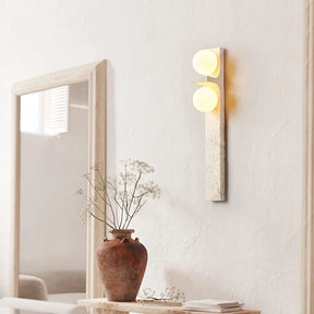 A modern Japan Style wall-mounted light fixture from Morsale.com, featuring two glowing circular LED bulbs, is vertically installed on a light-colored wall. Below the Natural Travertine Modern Wall Lights, to the left, a green leafy plant is partially visible.