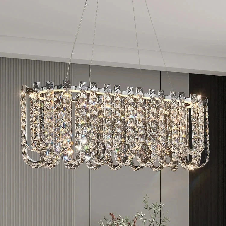 The Bacci Crystal Beads Chandelier by Morsale is a contemporary rectangular lighting fixture, showcasing exquisite craftsmanship with dangling crystal prisms that create a luxurious radiance. It hangs elegantly from the ceiling in a room decorated with light gray walls featuring vertical stripes.