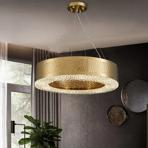 In a modern style living room, the Alleri Hammered Gold & Crystal Chandelier by Morsale with LED bulbs dazzles from the ceiling. Sheer curtains elegantly veil the window in the background, beautifully complementing a large green plant and a round wall mirror for a contemporary flair.