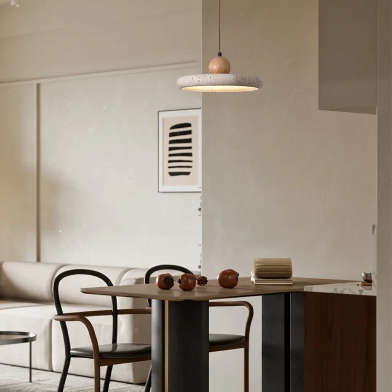 A minimalist dining area featuring a small table with dark legs and a light wooden top, surrounded by four chairs. The beige walls are adorned with a black-and-white abstract framed picture. Overhead, modern Natural Travertine Kitchen Pendant Light Fixtures from Morsale.com fitted with LED bulbs illuminate the space, which is subtly decorated with small items.