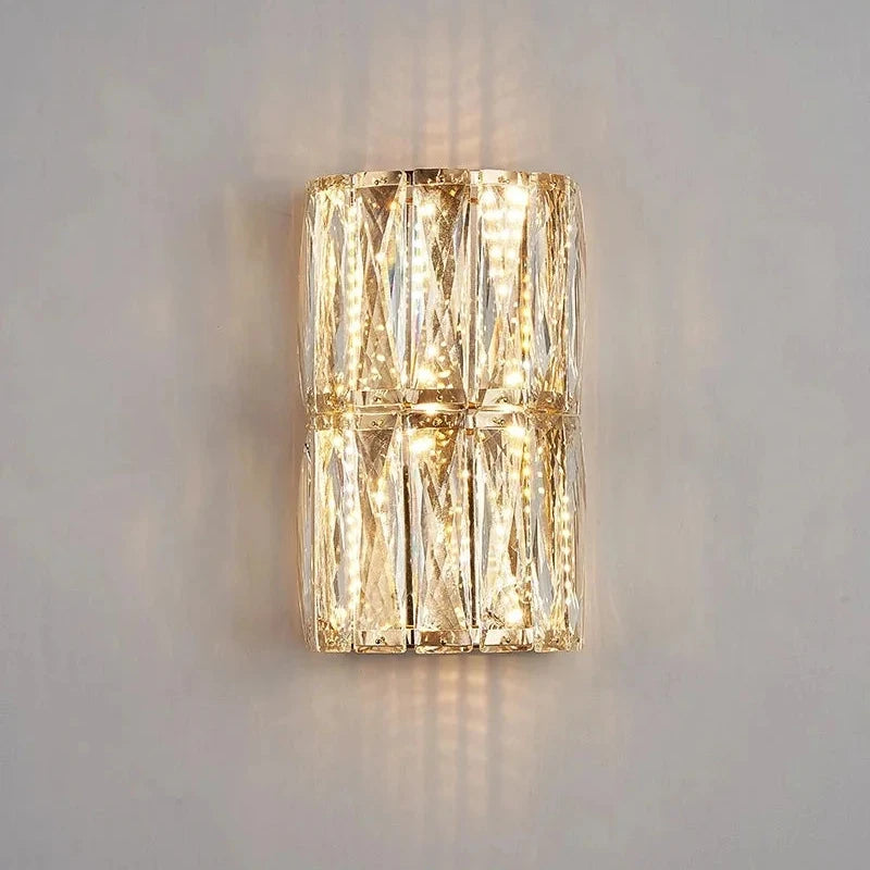 The Bacci Crystal Wall Lights from Morsale.com feature a modern, rectangular, transparent glass design that emits a warm white light. Adorned with handmade crystals arranged in a geometric pattern, these wall sconces are mounted on a plain white wall to create a soft ambiance.