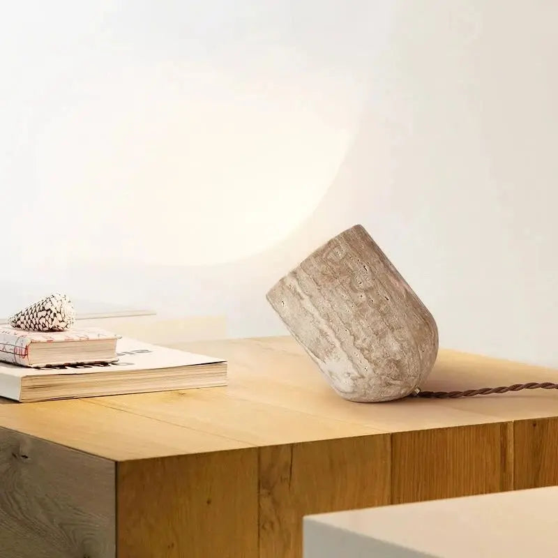 A small, cylindrical Modern Travertine Table Lamp from Morsale.com, with a concrete texture, tilts at an angle on a light-colored surface. Emitting a warm yellow light reminiscent of natural yellow cave stone, it casts a soft glow on the wall behind it. The power cord is visible trailing off to the side.