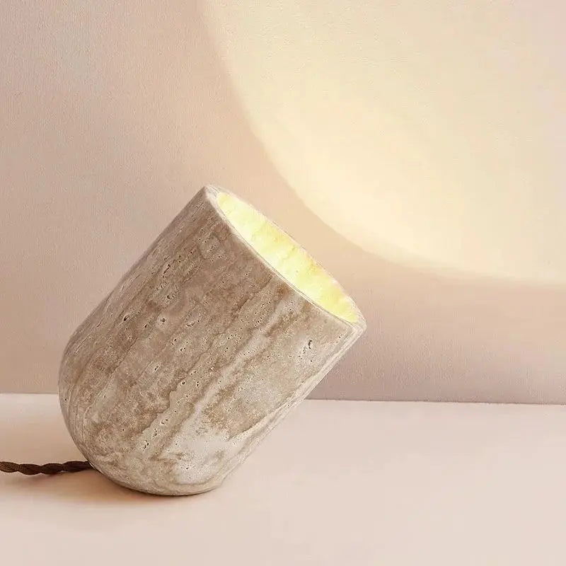 A small, cylindrical Modern Travertine Table Lamp from Morsale.com, with a concrete texture, tilts at an angle on a light-colored surface. Emitting a warm yellow light reminiscent of natural yellow cave stone, it casts a soft glow on the wall behind it. The power cord is visible trailing off to the side.