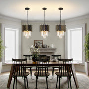 A dining room showcases modern style with a wooden table, six black chairs on a patterned rug, and three Gio Crystal Dining Area Light Fixtures by Morsale. Two large windows flank a fireplace adorned with decor, while potted plants grace either side of the space.