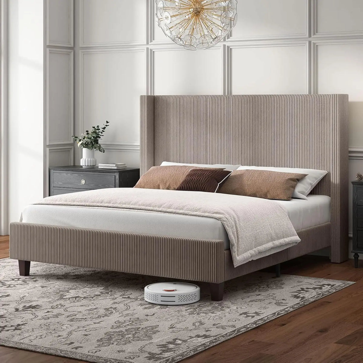 A modern bedroom features Morsale's Corduroy Bed with a wingback headboard in beige, paired with matching bedding. Two dark bedside tables sit by the bed, and a patterned rug covers the floor. Nearby, a white robotic vacuum rests under the chandelier's glow.